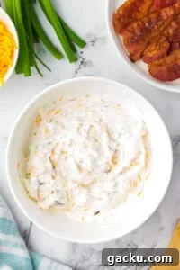Creamy Loaded Baked Potato Dip Recipe for Parties and Game Day 11 A white bowl filled with a sour cream based dip, with bowls of cooked bacon and cheese on the side.