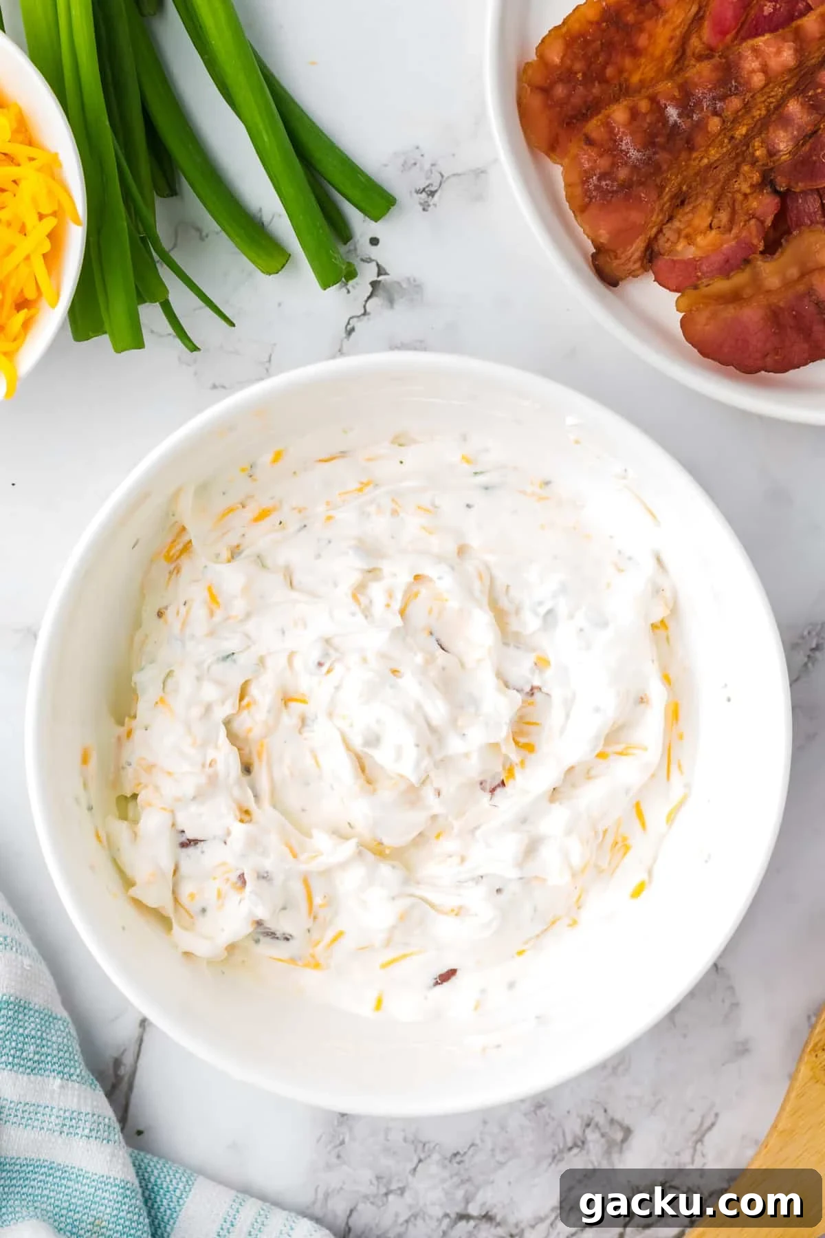 Creamy Loaded Baked Potato Dip Recipe for Parties and Game Day 5 A white bowl filled with a sour cream based dip, with bowls of cooked bacon and cheese on the side.