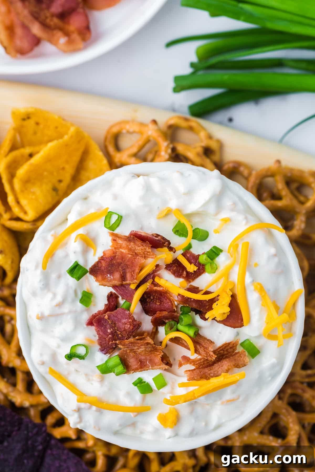 Creamy Loaded Baked Potato Dip Recipe for Parties and Game Day 6 An overhead shot of a bowl of cream dip garnished with green onion, shredded cheese, and bacon. Surrounded by corn chips and pretzels.