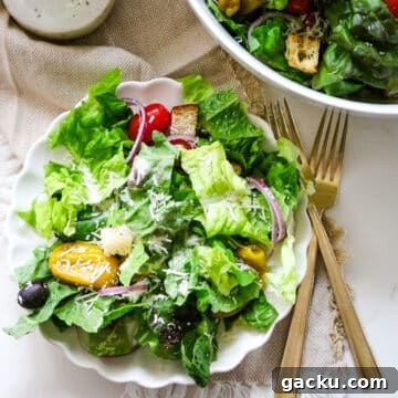 A fresh green salad on a white plate with tomatoes, onions, olives, and croutons, plus two gold forks beside it.