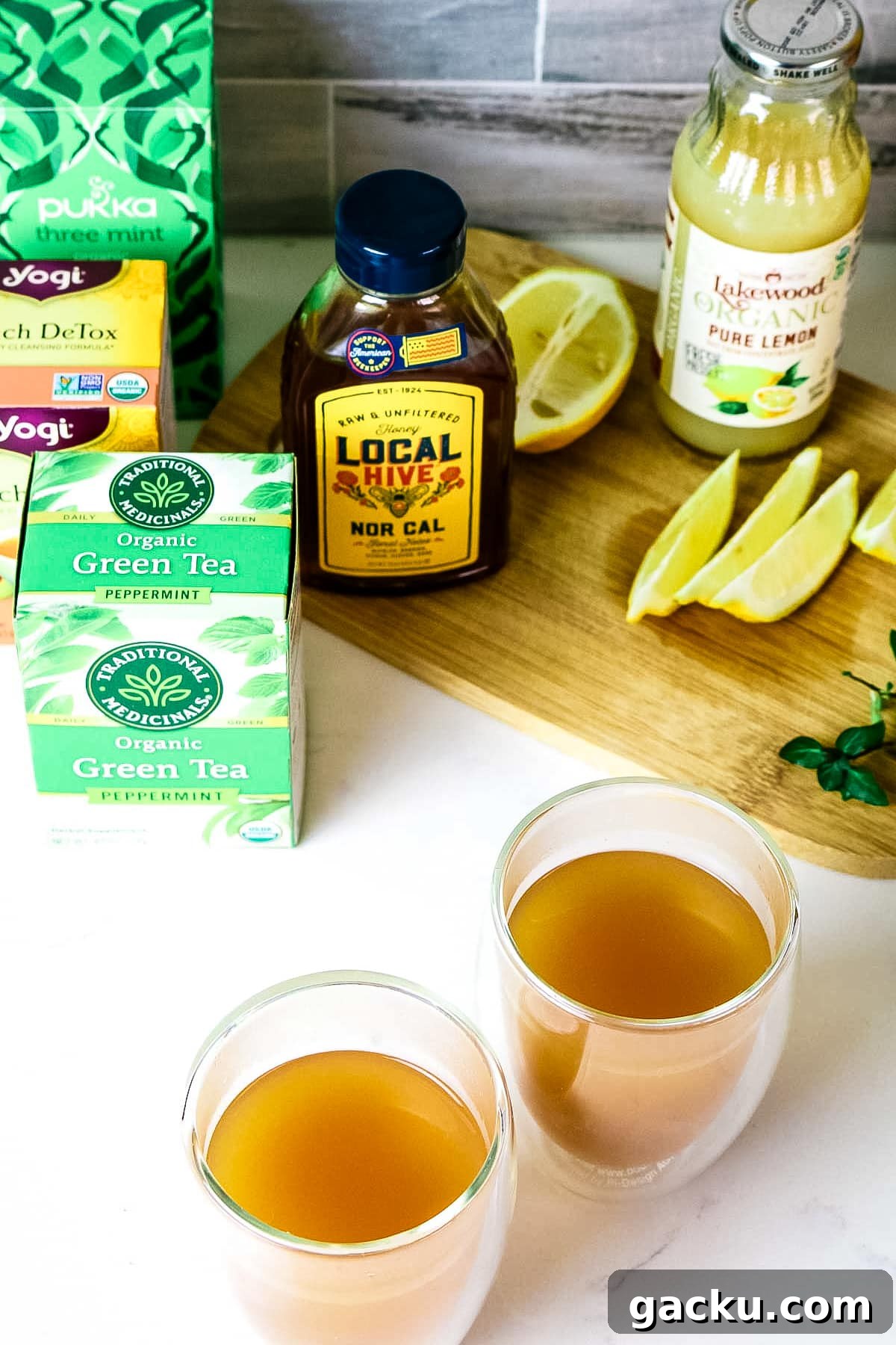 two clear glasses with herbal tea, tea boxes, lemon juice, and raw honey bottle.