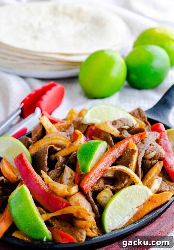 Steak fajitas in fajita skillet with peppers, lime wedges, and tortillas. 
