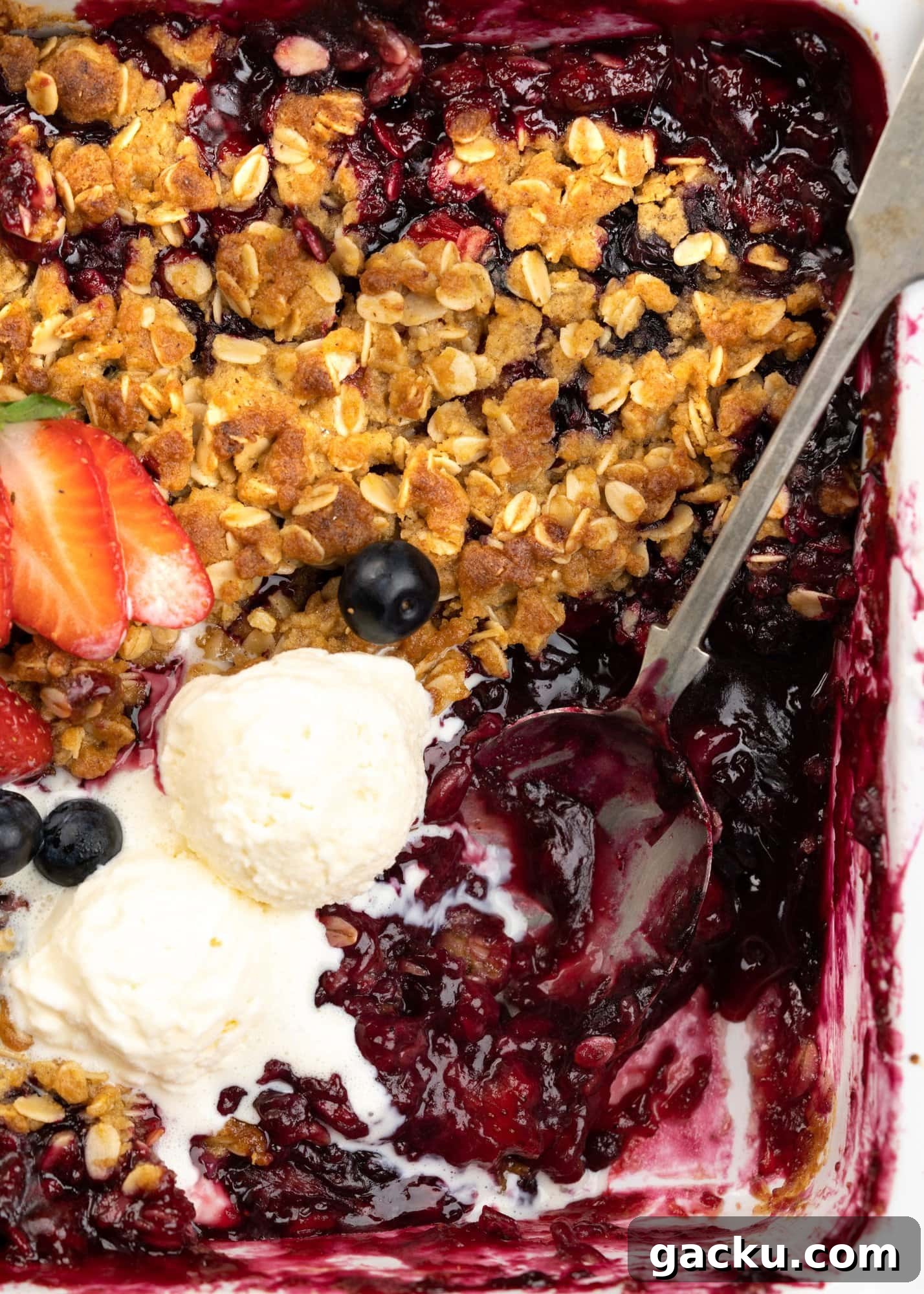 A corner of baking tray shows two generous scoops of vanilla ice cream on a golden brown crisp oatmeal layer and underneath is a gooey berry filling. 