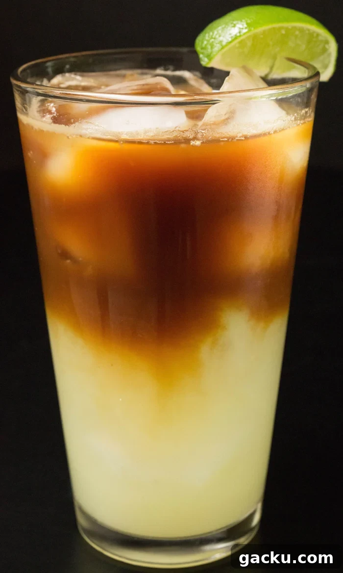 A layered drink in a tall glass with ice and a lime wedge. Green lime juice bottom layer and brown coffee top layer.