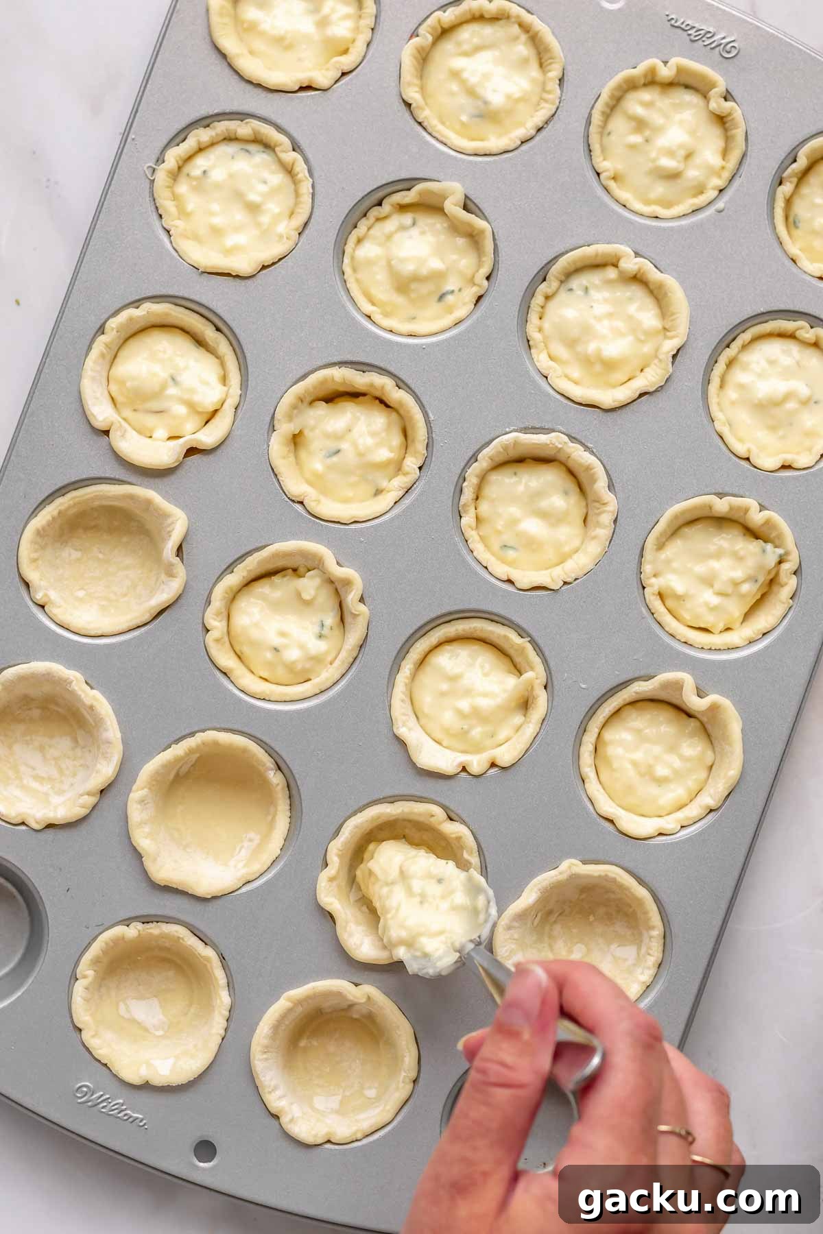Mini Cheese Tartlets Recipe: Crispy Crusts with Creamy Filling 9 A spoon adds cheese filling into puff pastry shells in a mini muffin tin.