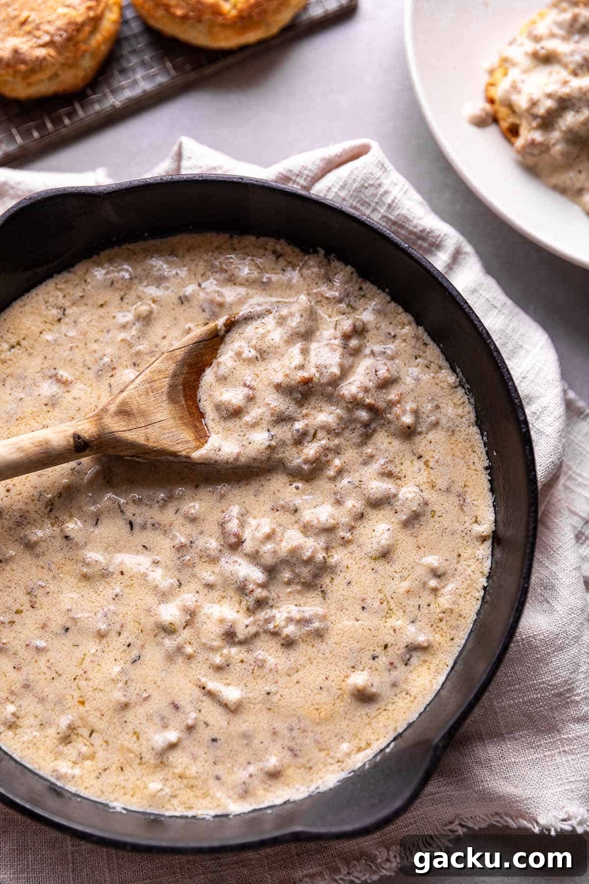 Southern-Style Sausage Gravy Recipe for Biscuits and Breakfast 2 A cast iron pan with a sausage grave and wooden spoon with a plate of biscuits and gravy in the back.