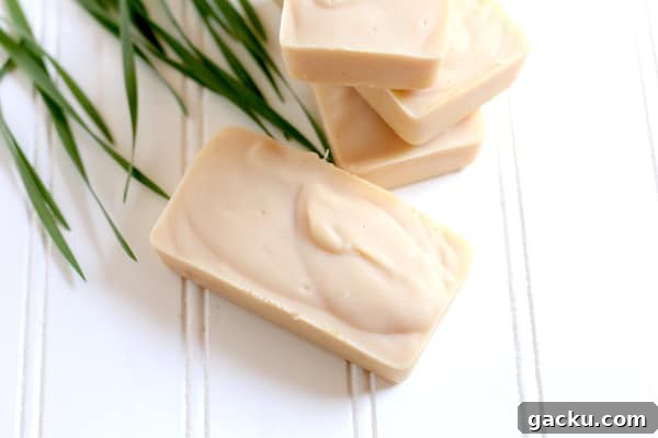 Image shows four small bars of lemongrass goat milk soap on white shiplap, sitting next to blades of fresh lemongrass.