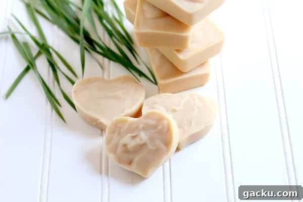 Photo shows three bars of heart shaped lemongrass soap on a white shiplap board in front of a stack of rectangle bars of soap and blades of lemongrass.