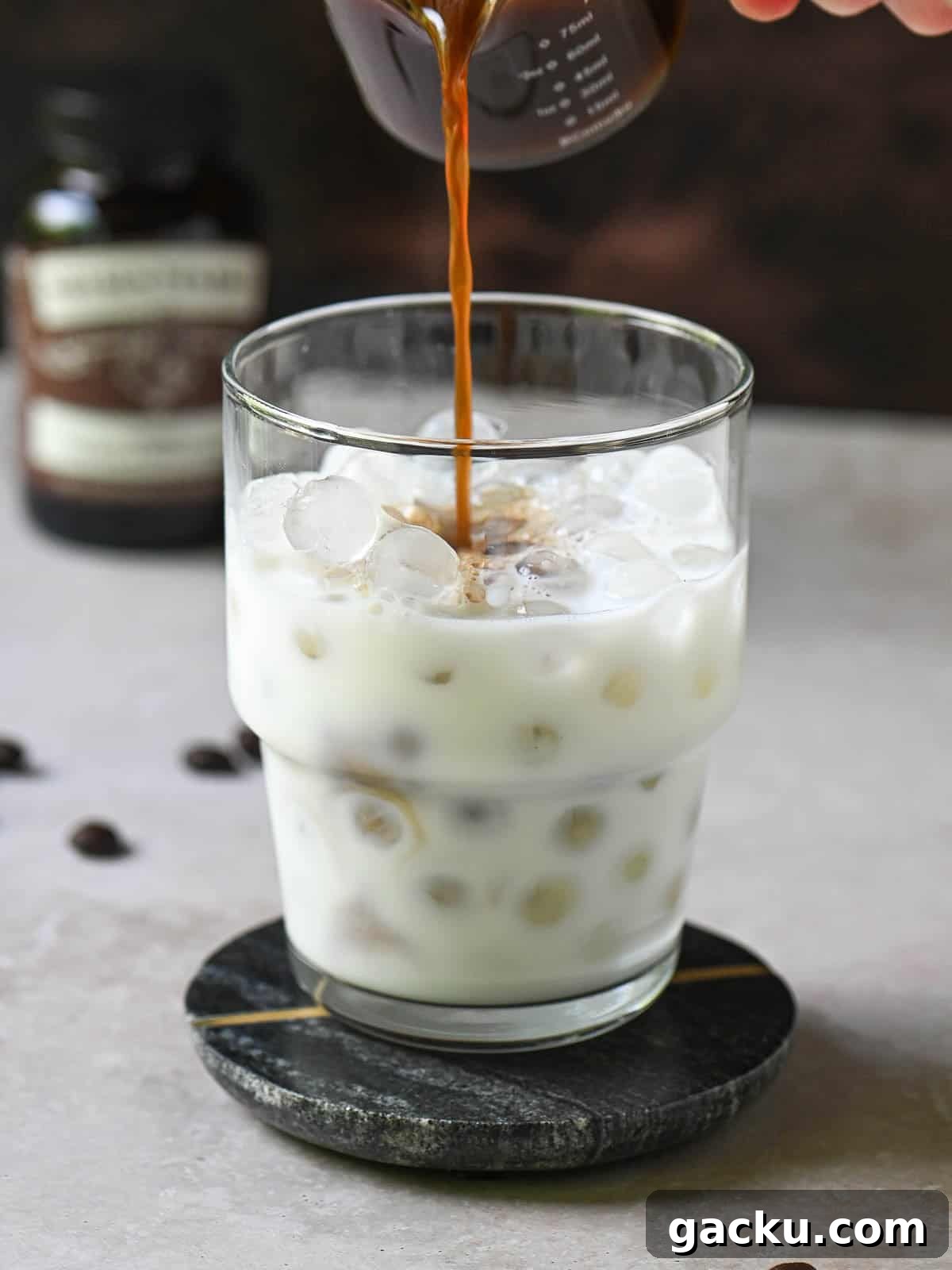 Coffee is being poured into a clear glass with milk and ice.