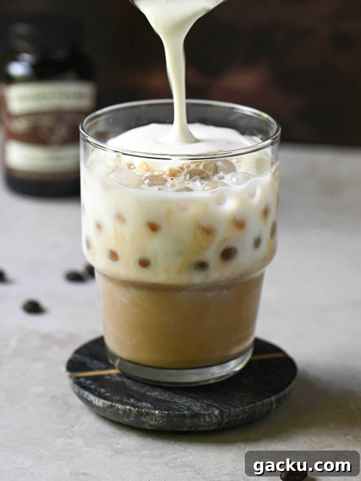Cold foam is being poured into a clear glass with coffee and milk.