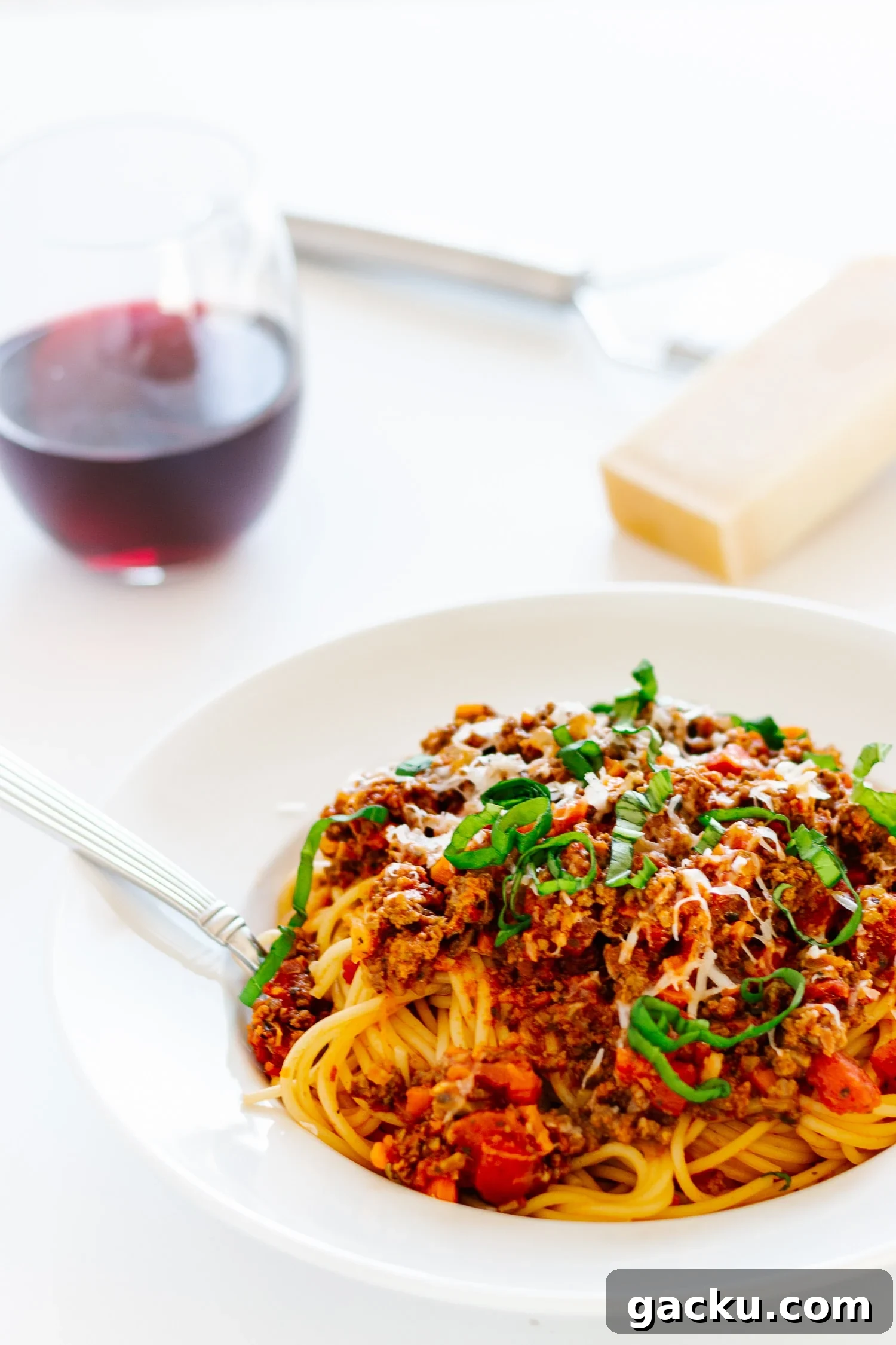 Authentic Bolognese Sauce Recipe for Rich, Slow-Simmered Pasta 5 White bowl of spaghetti bolognese with basil scattered on top and a glass of red wine and chunk of parmesan and a grater in the background
