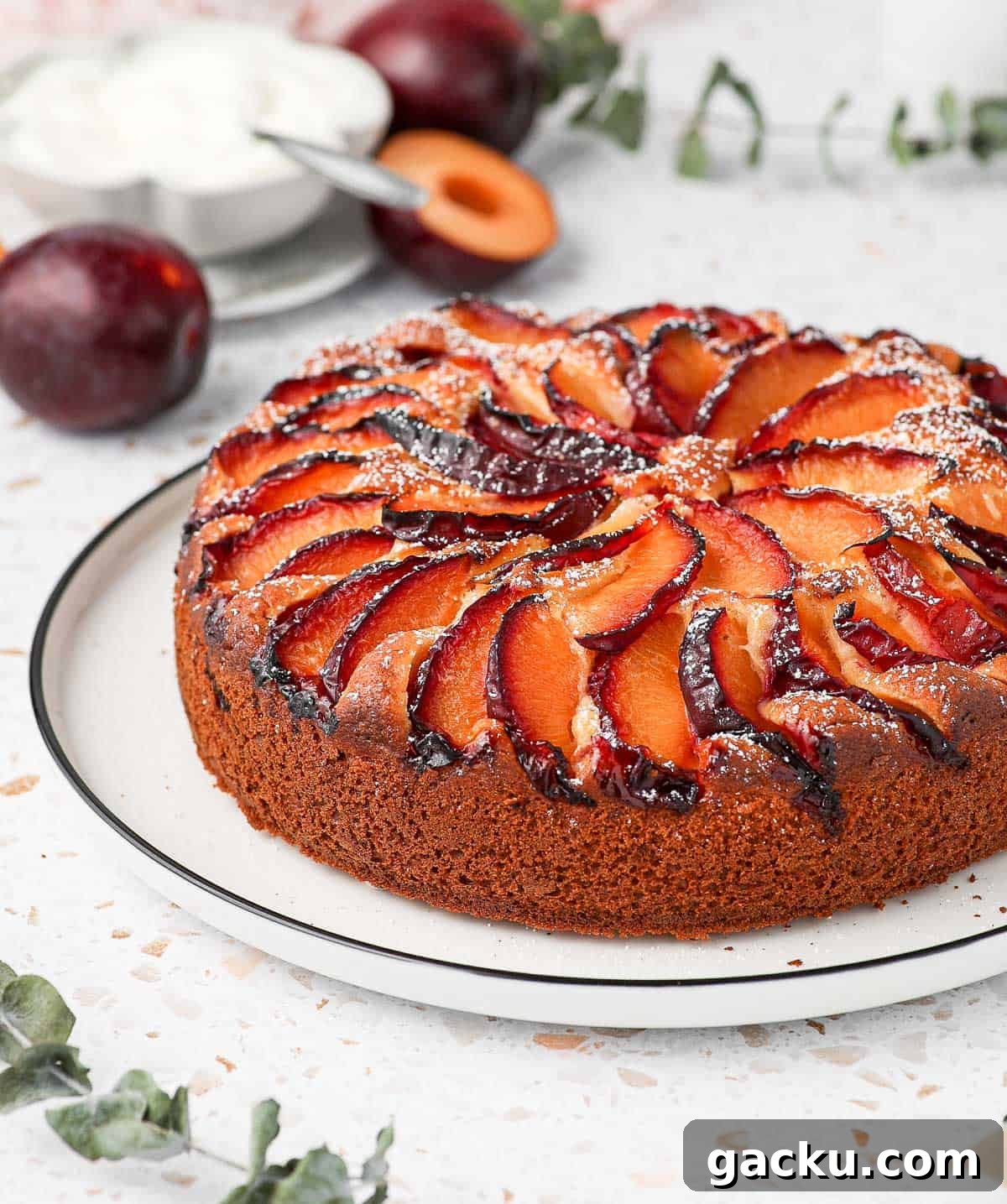 Plum Cake on a white plate surrounded by leaves, plums and a bowl of yogurt.