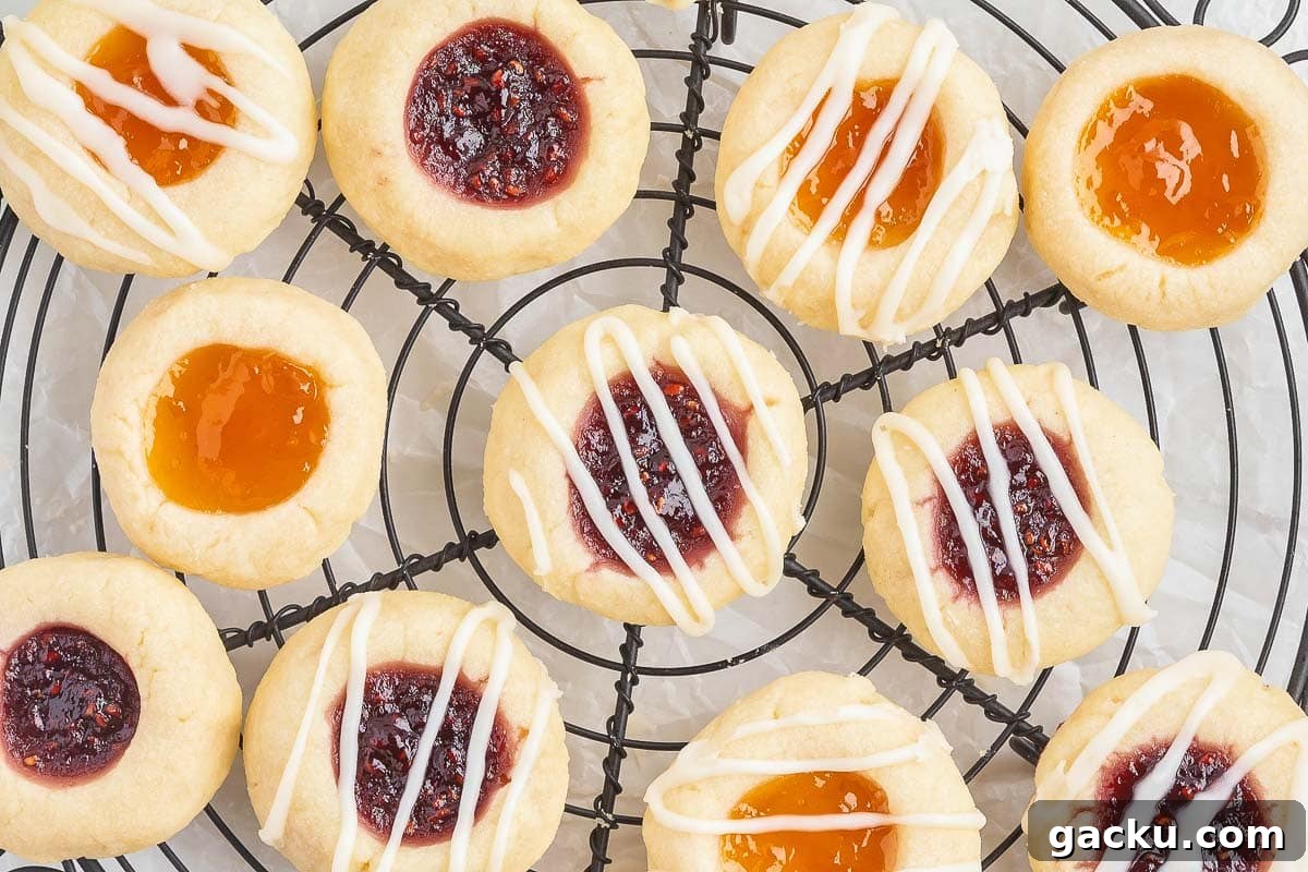 A cooling rack holds an assortment of round thumbprint cookies with various fillings, including red and orange jams and white icing drizzles.