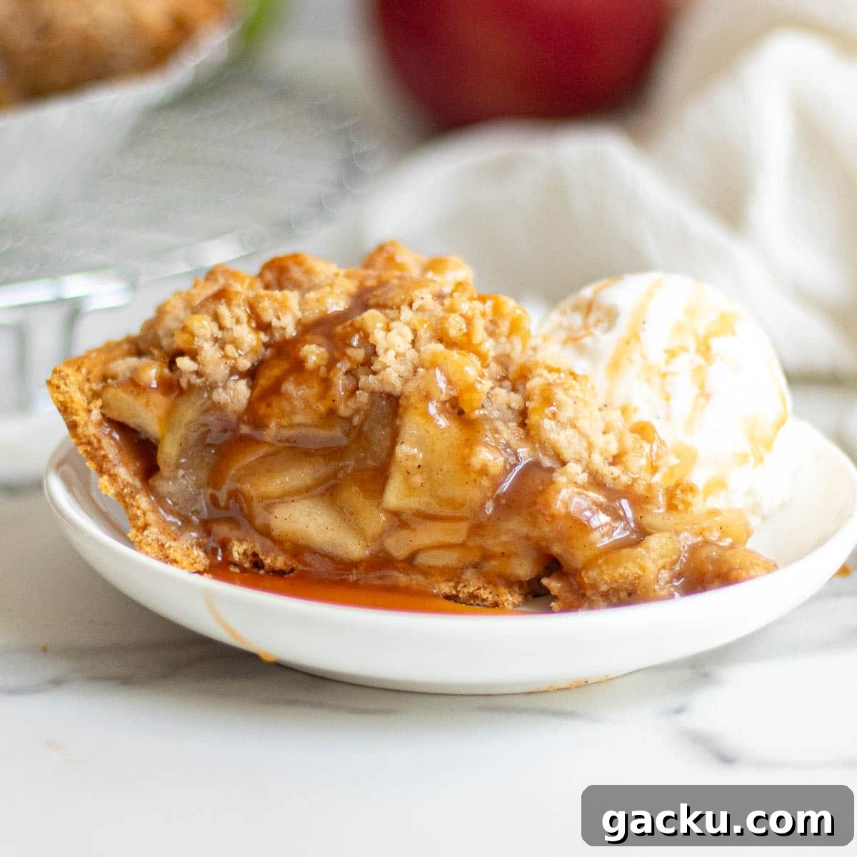 A slice of apple pie in a graham cracker crust drizzled with caramel sauce and served with a scoop of vanilla ice cream on a white plate.