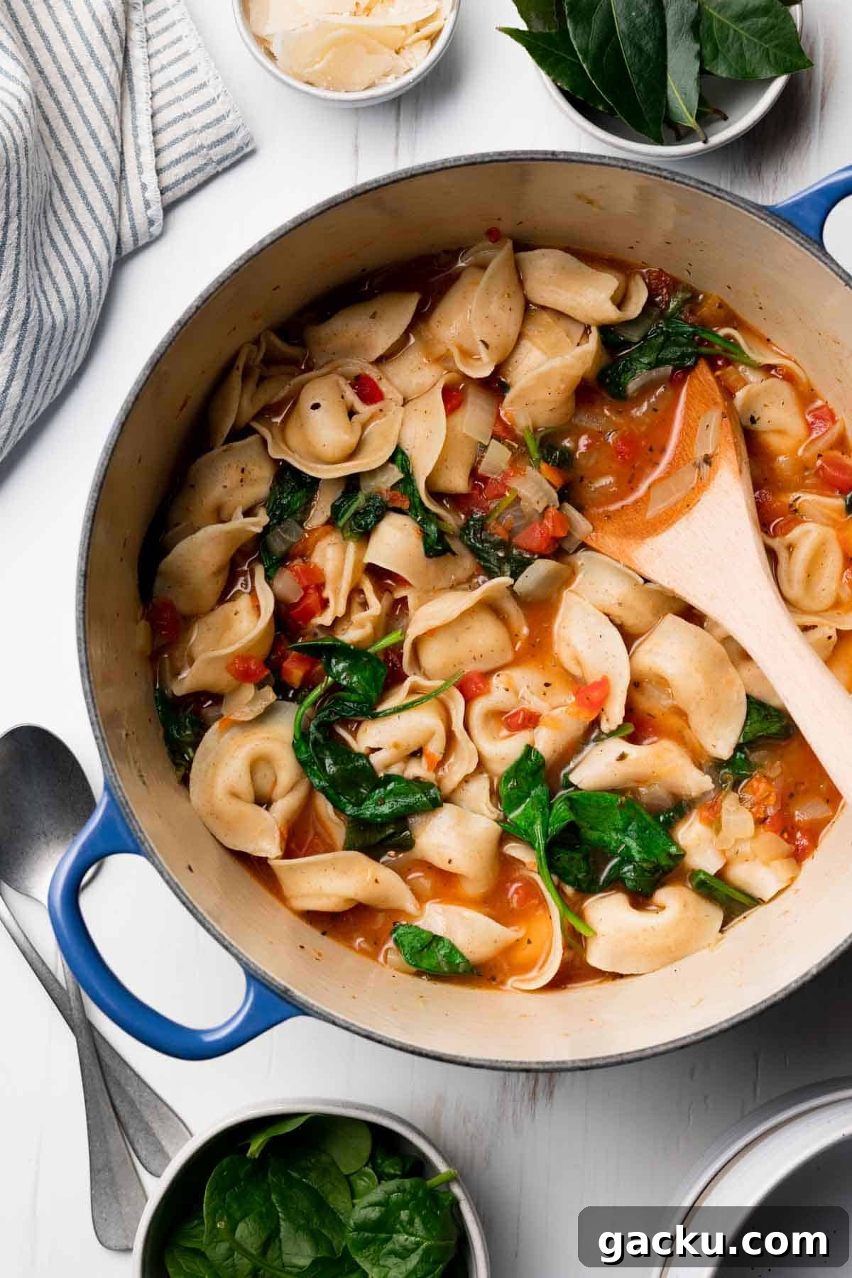 Gluten free spinach tortellini soup in large blue pot next to baby spinach and shaved parmesan cheese.