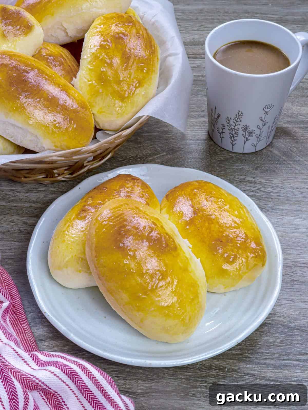 Yema Bread Rolls Recipe — Soft Filipino Custard-Filled Buns 2 Yema Bread Rolls on a white plate with a cup of coffee and a basket of rolls in the background.