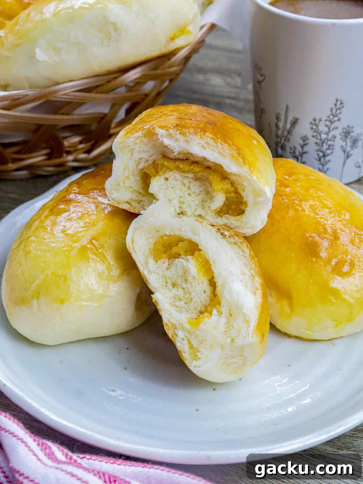 Yema Bread Rolls Recipe — Soft Filipino Custard-Filled Buns 7 Filipino custard buns on a white plate with a basket of bread rolls and cup of coffee in the background.