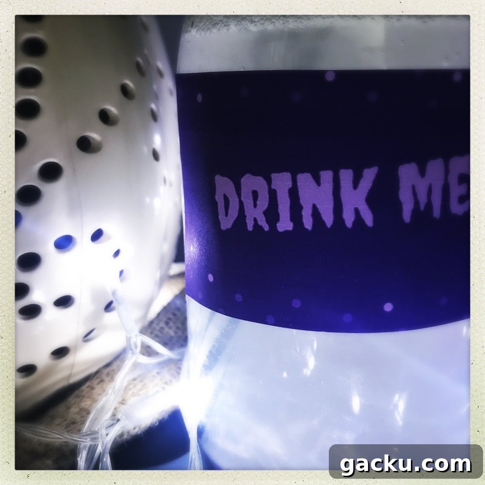 bottle of silver Halloween mocktail drink with label on saying Drink Me, white pumpkin in background.