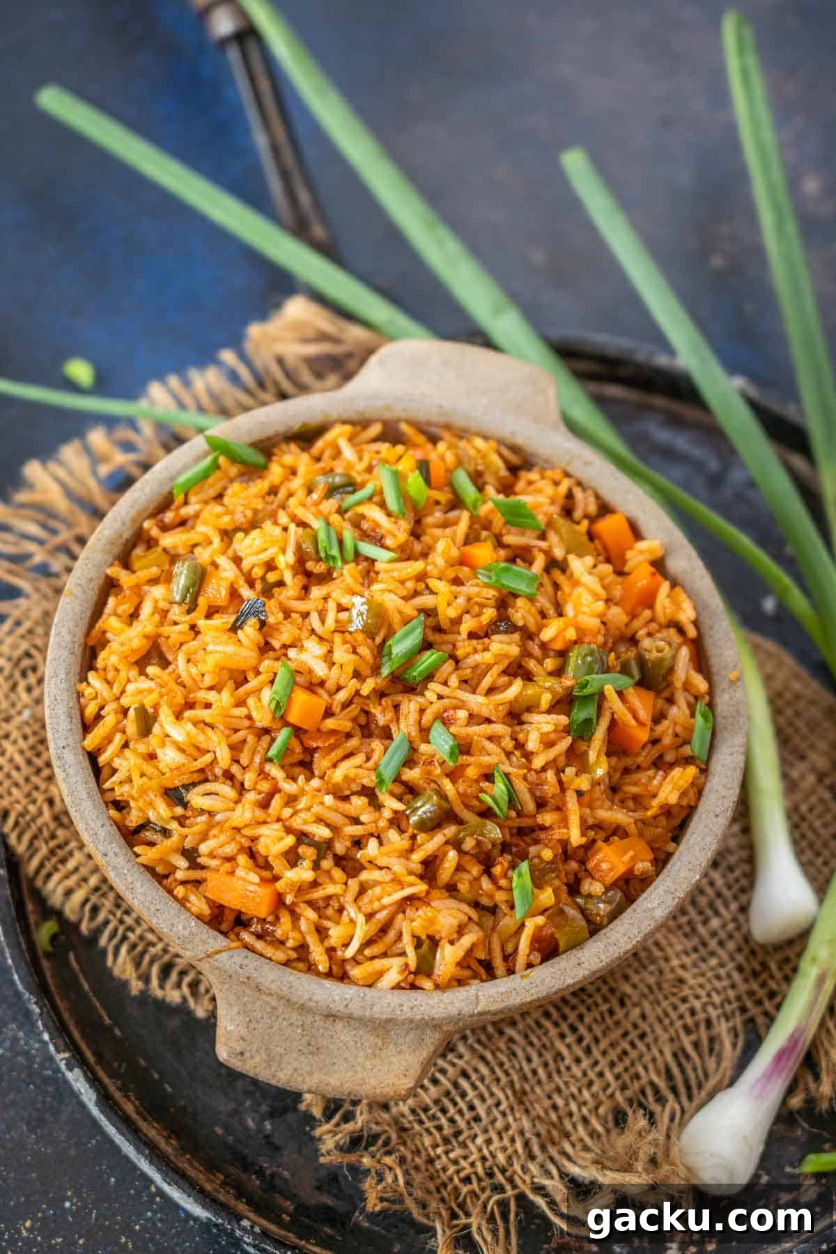 A bowl of Schezwan Fried Rice garnished with chopped green onions sits on burlap, with fresh spring onions beside it, all set on a dark rustic surface.