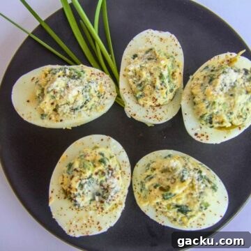 Keto Spinach and Artichoke Deviled Eggs Recipe 5 Five keto deviled eggs rest on a black plate, garnished with spices and green herbs, next to fresh chives.