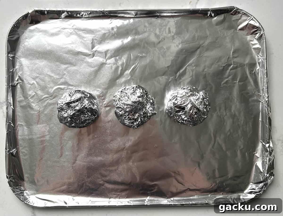 Roasted Garlic Butter Recipe for Homemade Bread, Steak & Veggies 4 Three garlic heads wrapped in foil parcels on a baking sheet.