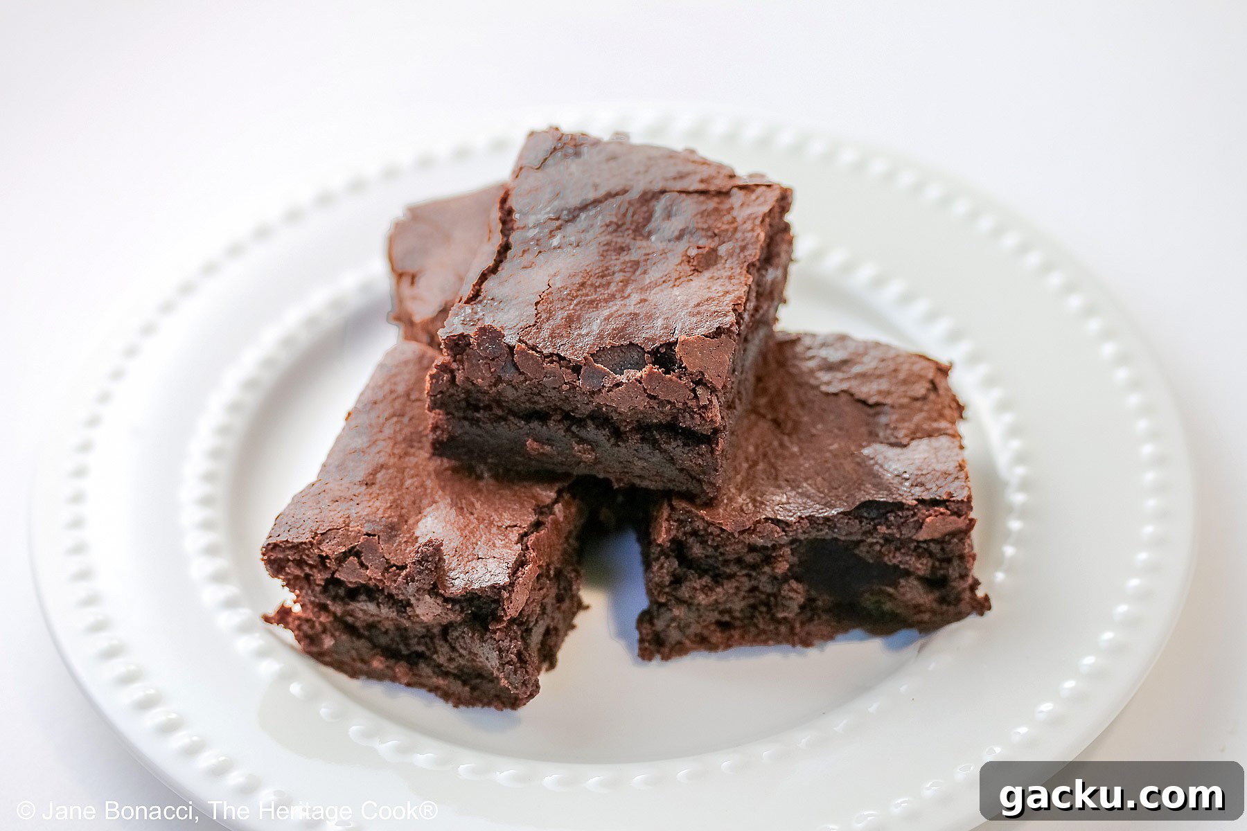 The Best Chewy Brownies, rich and so chocolatey, stacked on a white plate with two rows of white pearls, sitting on a white background; some shots of the cookbook, “Bittersweet” by Alice Medrich; © 2025 Jane Bonacci, The Heritage Cook.
