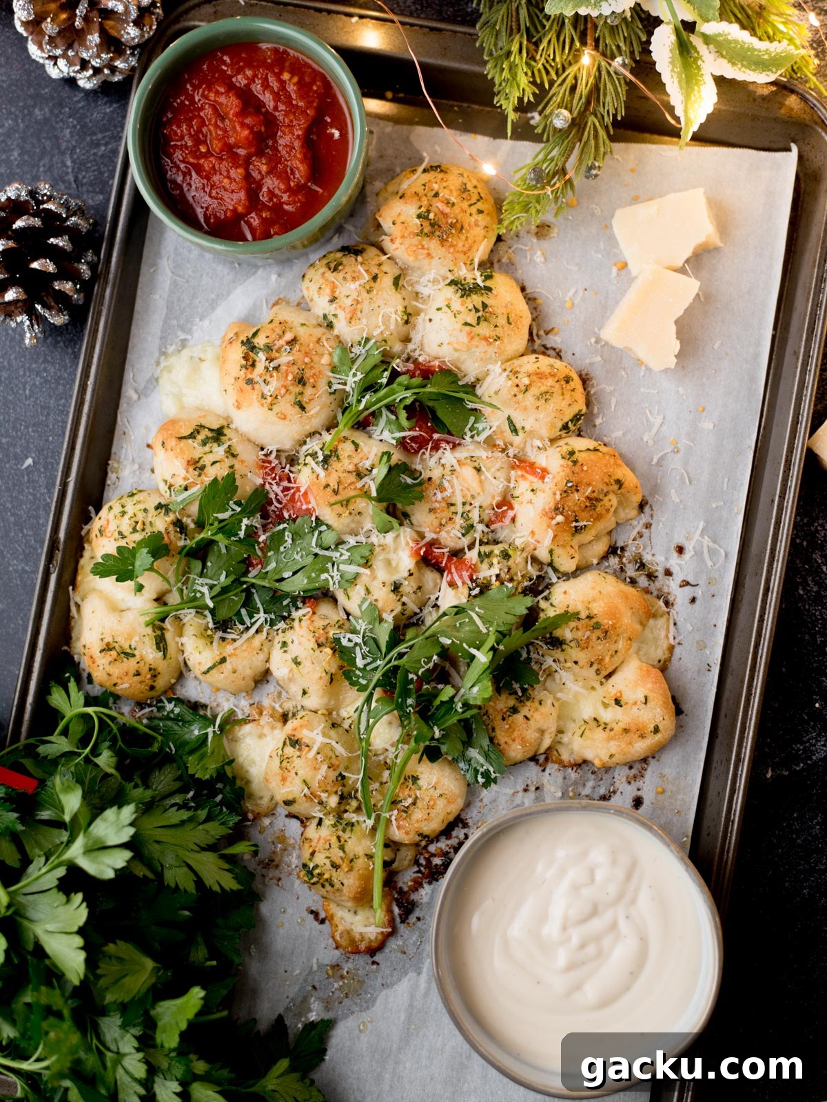 Christmas Tree Pull-Apart Bread Recipe for Holiday Brunch 2 A festive, tree-shaped pull-apart bread with herbs and cheese on a baking tray, served with tomato sauce and a creamy dip, garnished with fresh parsley and Parmesan cheese. Holiday decor surrounds the tray.