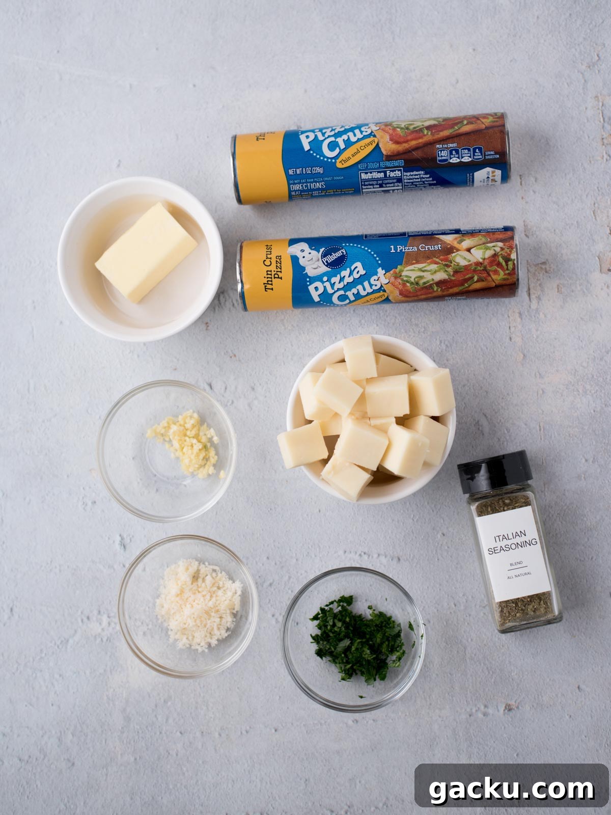 Christmas Tree Pull-Apart Bread Recipe for Holiday Brunch 3 Ingredients on a light surface: two cans of pizza crust, a bowl of butter, cubed cheese, grated cheese, minced garlic, chopped herbs, and a jar labeled Italian Seasoning.