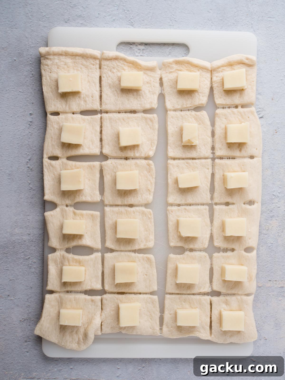Christmas Tree Pull-Apart Bread Recipe for Holiday Brunch 4 A white cutting board with rectangular dough pieces, each topped with a small square of cheese, arranged in a grid pattern on a light gray surface.