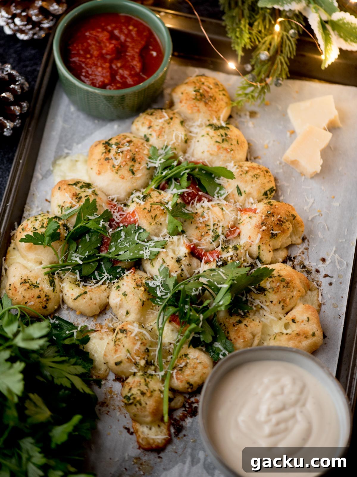 Christmas Tree Pull-Apart Bread Recipe for Holiday Brunch 7 A tray of pull-apart garlic bread bites topped with herbs and grated cheese, garnished with fresh arugula, served with bowls of marinara sauce and creamy dip, and pieces of parmesan on the side.