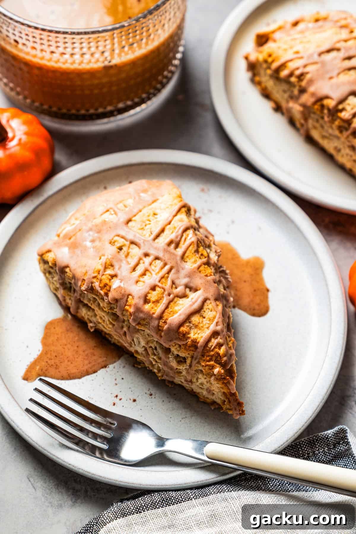 These tender, flaky cinnamon scones are packed with wholesome oats and cozy fall spice! A glazed cinnamon scone next to a fork on a plate, with a second scone and a cup of tea in the background.