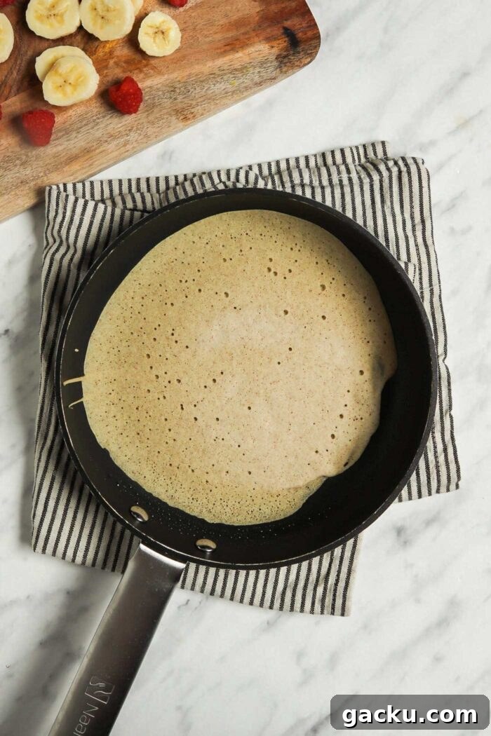 A buckwheat crepe cooking in a skillet with chopped strawberries and banana beside it.