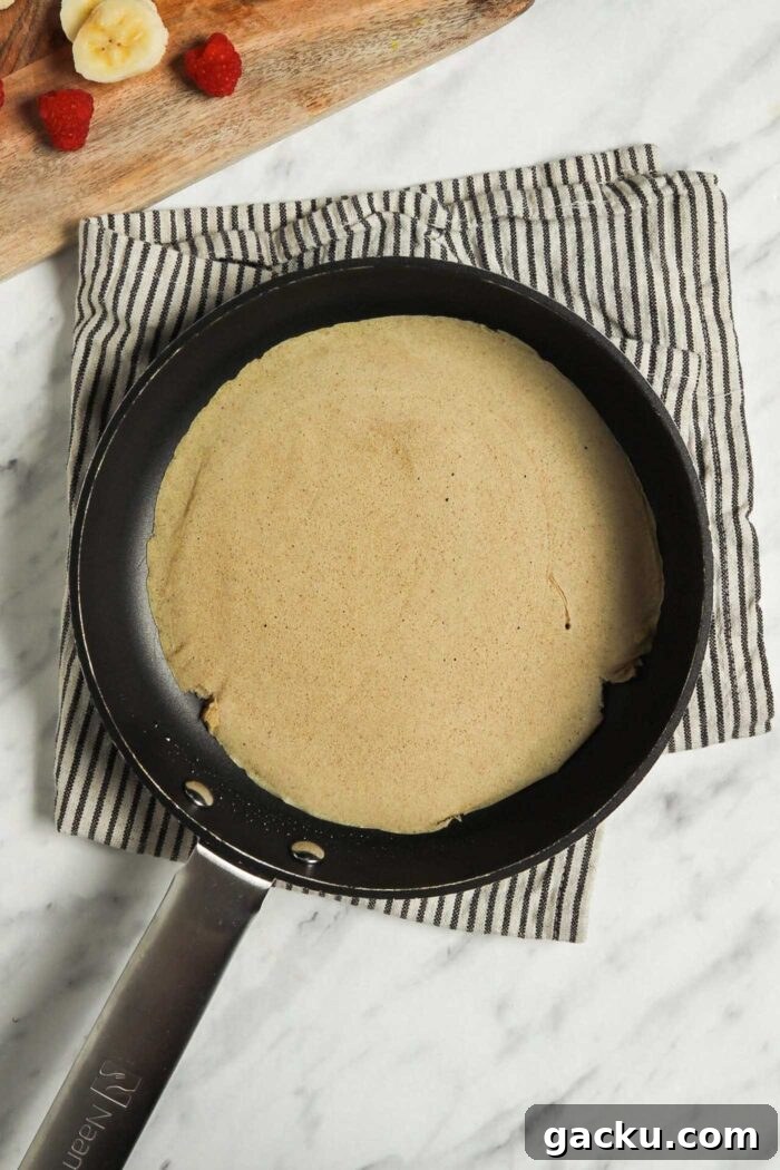 Vegan buckwheat crepe in a skillet with a cutting board beside it with chopped strawberries and banana on it.