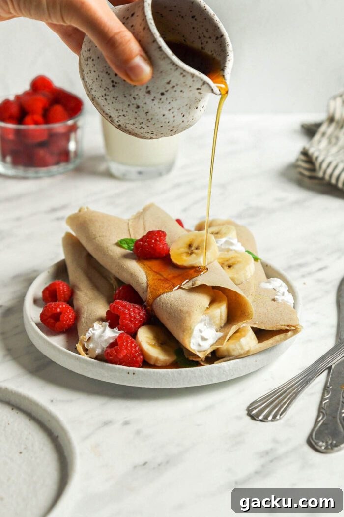 Hand pouring maple syrup from a small container over a stack of buckwheat crepes filling with strawberries and banana on a plate.