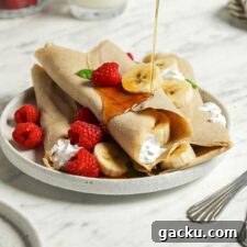 Pouring maple syrup from a small container over a stack of buckwheat crepes filling with strawberries and banana on a plate.