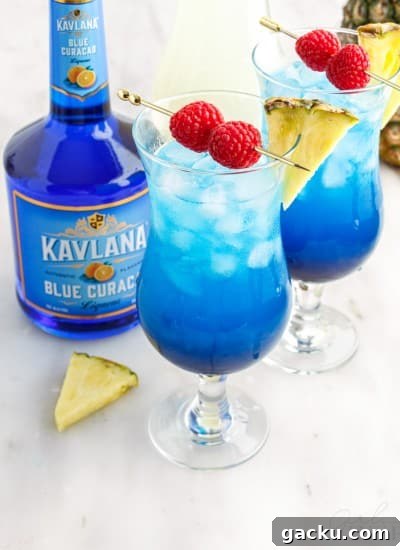 Close up of two tall glasses with fruit tingle cocktail and garnished with pineapple wedges and skewered raspberries, bottle of Blue Curacao, pineapple wedge garnish on a marble countertop.