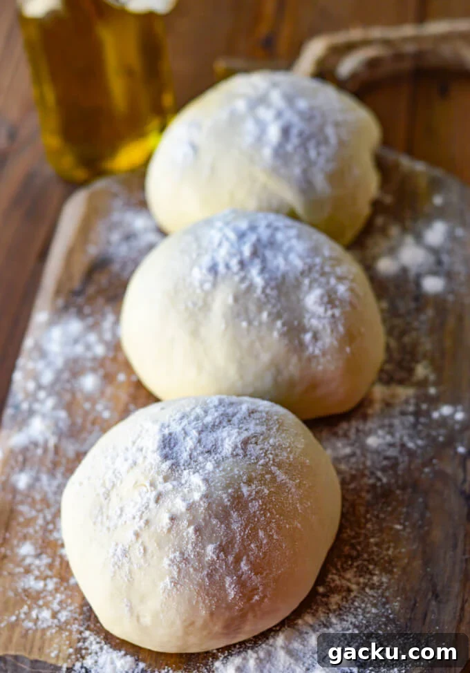 Three balls of pizza dough rest on a floured wooden board with olive oil in the background.