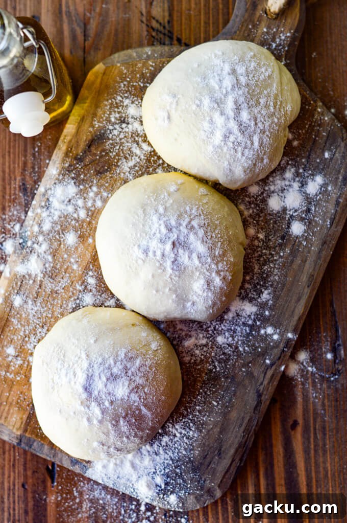 Three balls of pizza dough rest on a floured wooden board with olive oil in the background.