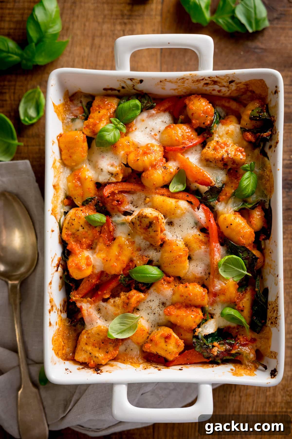 Creamy Baked Gnocchi with Chicken and Parmesan Crisp 2 Baked gnocchi with melted cheese, tomatoes, spinach, and fresh basil in a white oven dish. The dish is on a wooden table with a spoon and napkin beside it.