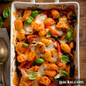 Creamy Baked Gnocchi with Chicken and Parmesan Crisp 12 Overhead square image of baked gnocchi with melted cheese, tomatoes, spinach, and fresh basil in a white oven dish. The dish is on a wooden table with a spoon and napkin beside it.