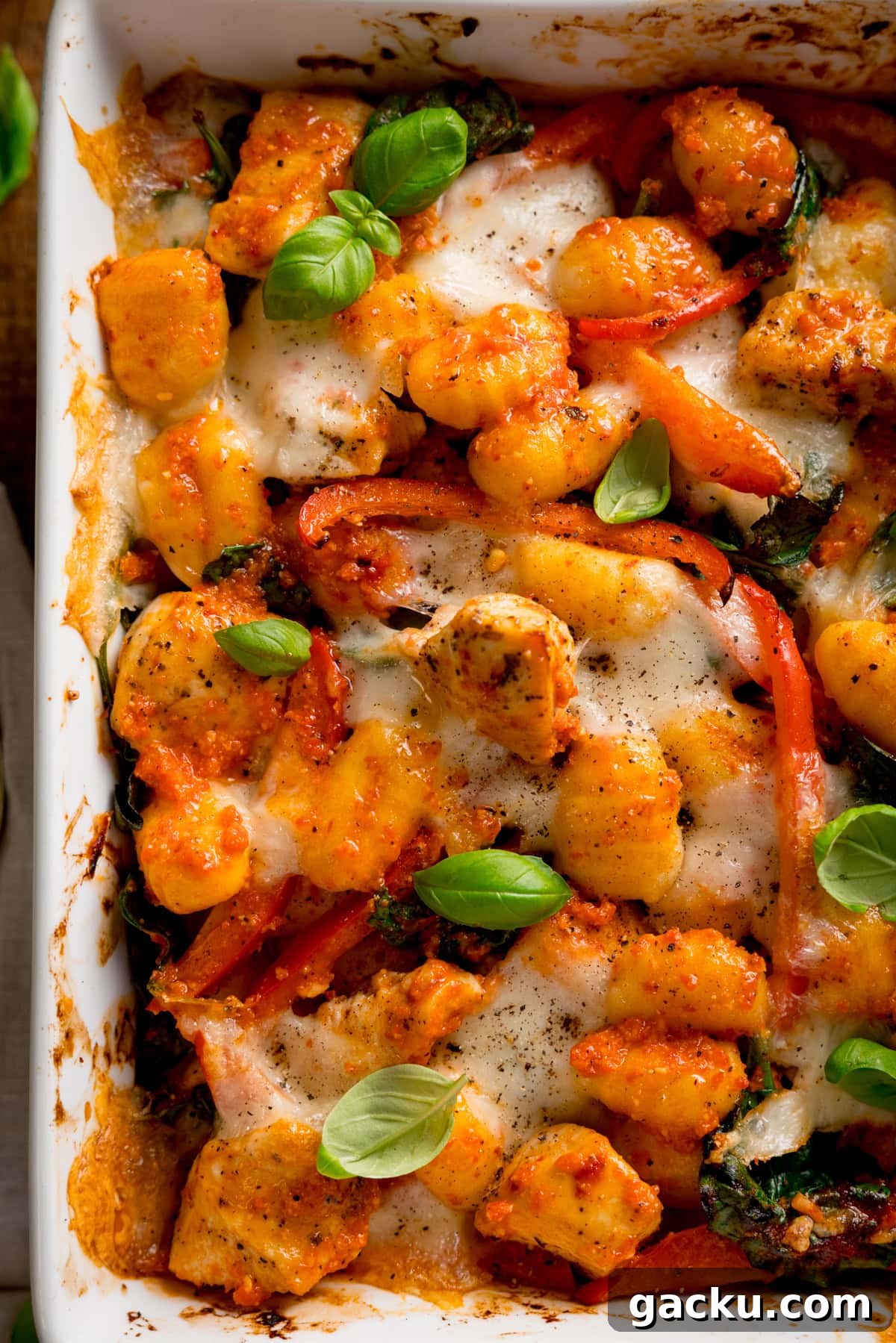 Creamy Baked Gnocchi with Chicken and Parmesan Crisp 4 Overhead close up of baked gnocchi with melted cheese, tomatoes, spinach, and fresh basil in a white oven dish.