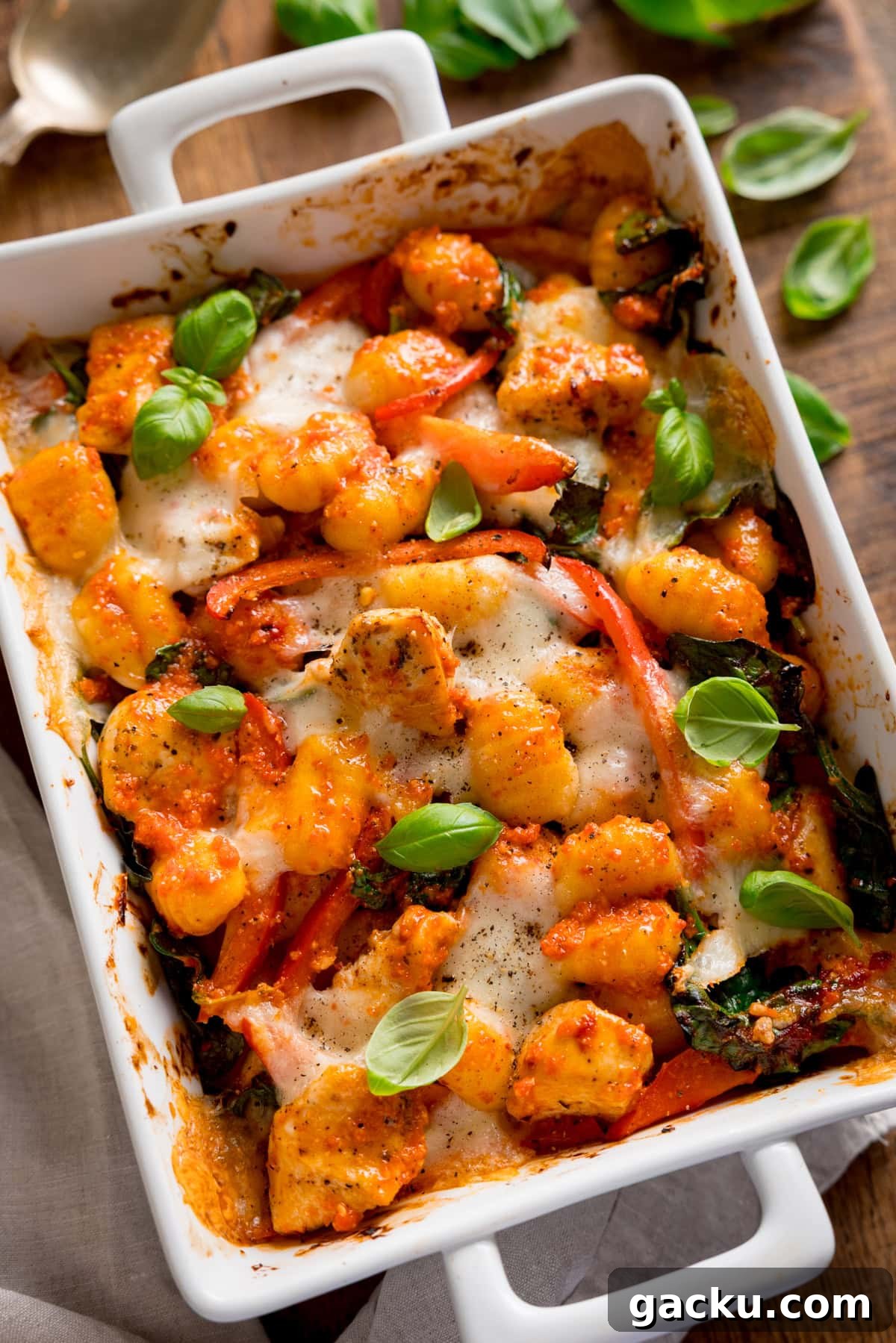 Creamy Baked Gnocchi with Chicken and Parmesan Crisp 5 Overhead image of baked gnocchi with melted cheese, tomatoes, spinach, and fresh basil in a white oven dish. The dish is on a wooden table with a scattering of basil leaves next to it.