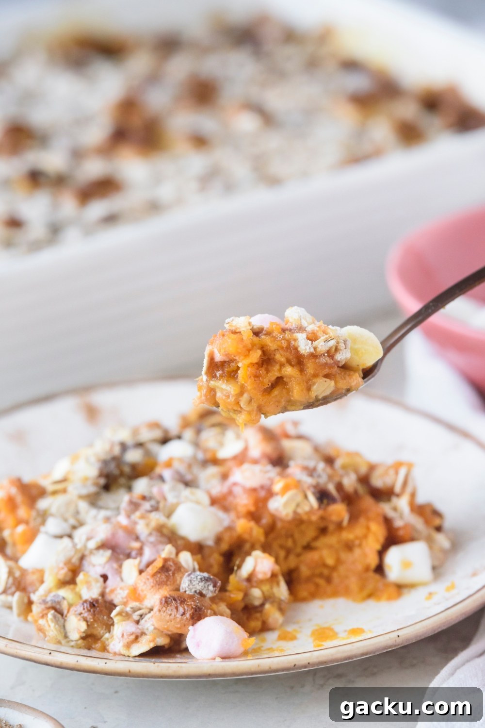 Someone lifting a spoonful of sweet potato casserole from a white plate. A full casserole dish is in the background.