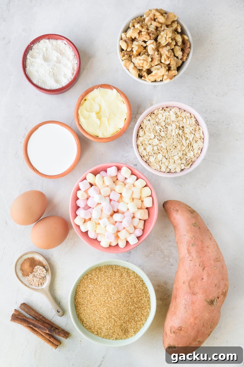 Walnuts, flour, cream, oats, mini marshmallows, butter, and brown sugar are in separate bowls. A large sweet potato is next to them on a white counter.