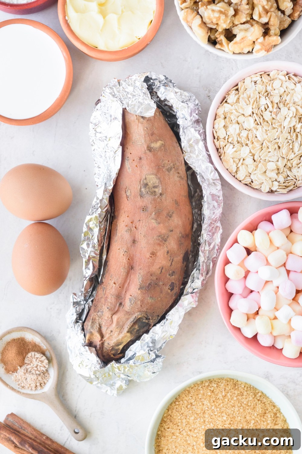 One large sweet potato partially wrapped in foil is on a white counter. Surrounding the potato are bowls of walnuts, oats, marshmallows, brown sugar, flour, cream, and two eggs.