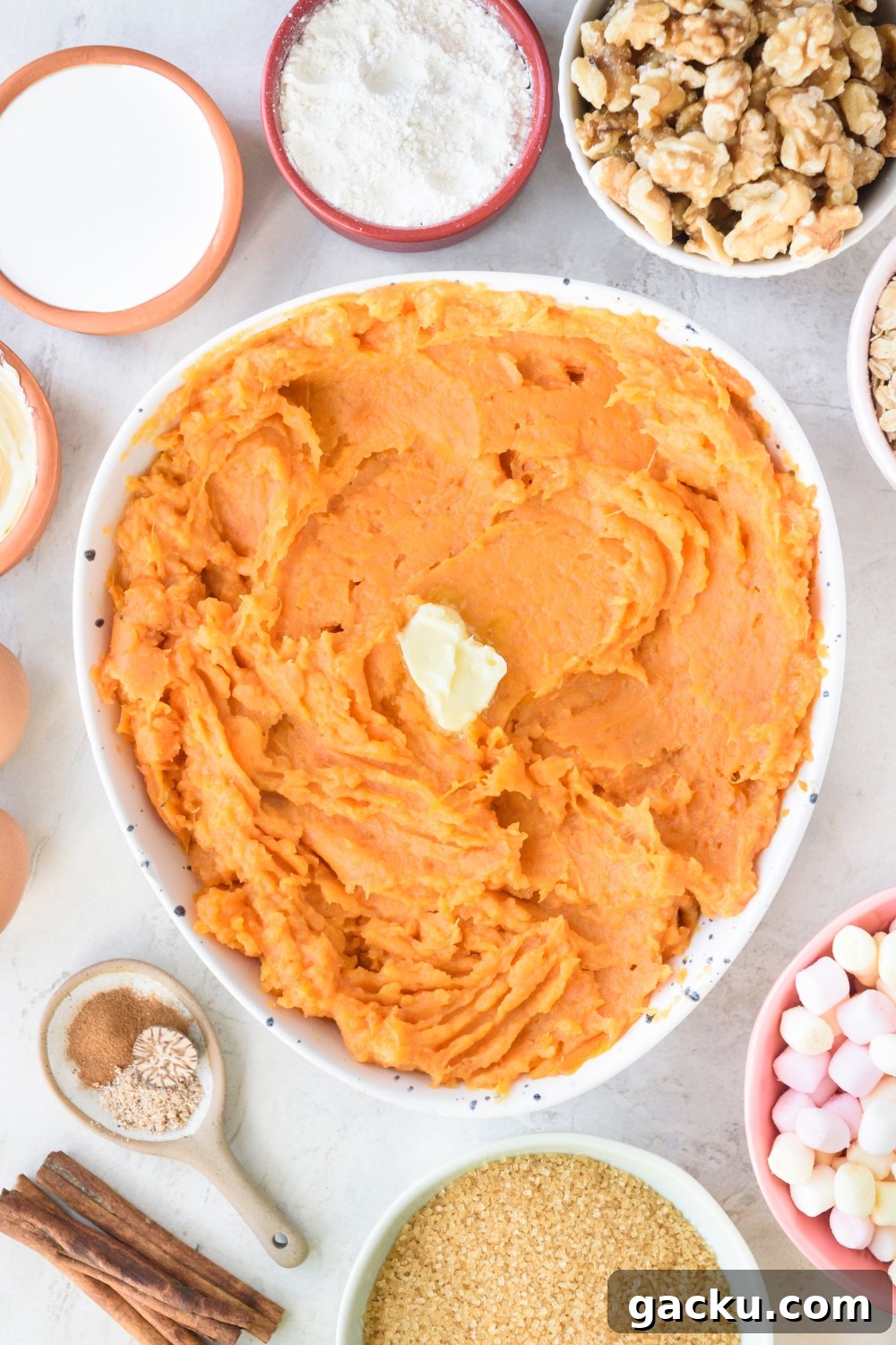 A large white bowl full of mashed sweet potatoes with a pat of butter in the middle. Bowls full of walnuts, marshmallows, brown sugar, spices, flour, and cream surround the large bowl