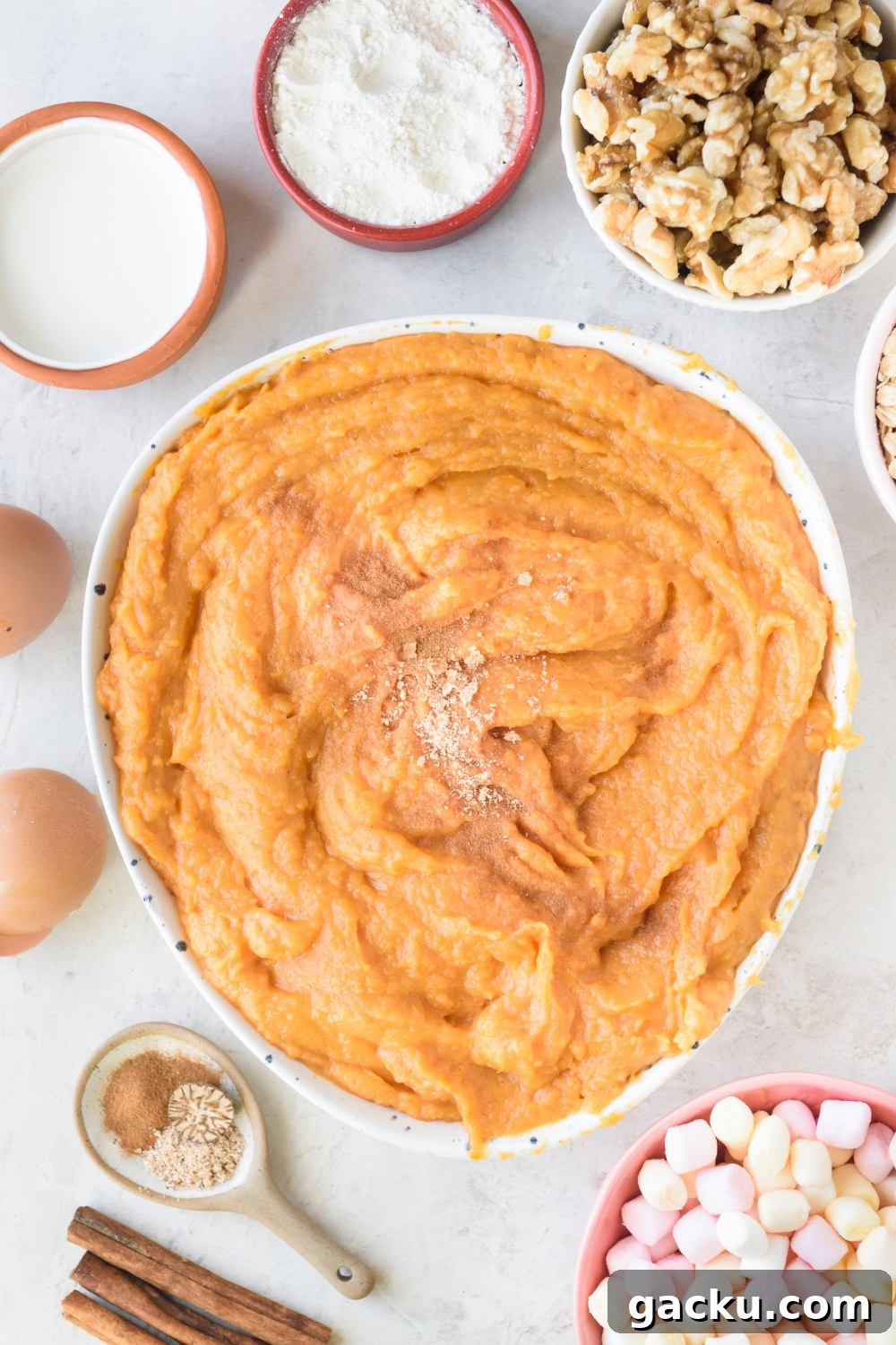 A large bowl of mashed sweet potatoes with spices on top on a white counter. Bowls of walnuts, flour, cream, 2 eggs, spices, and mini marshmallows surround the bowl.