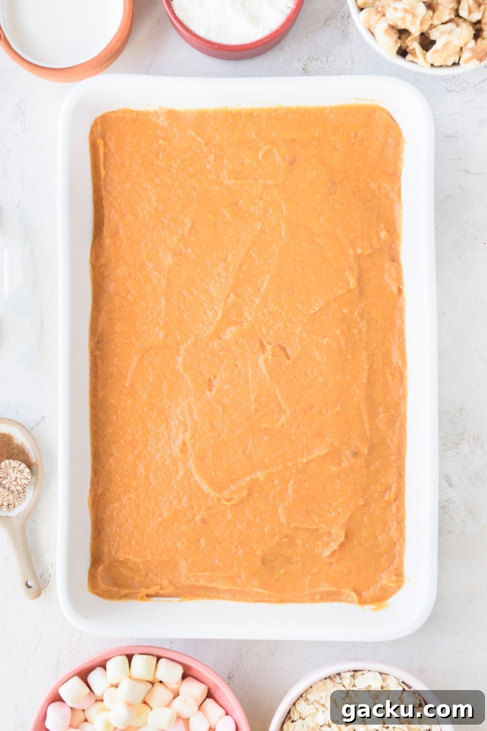 A white casserole dish is full of mashed sweet potatoes in an even layer. Bowls of marshmallows, oats, walnuts, flour, and cream surround the dish.
