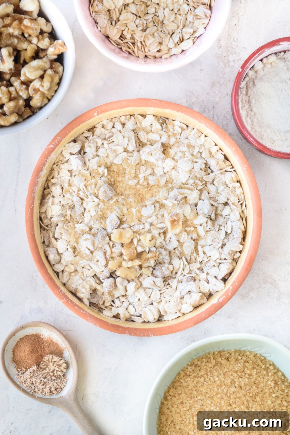 An orange bowl full of oats, walnuts, cinnamon, and sugar. Small bowls of walnuts, oats, flour, brown sugar, and spices surround the bowl.