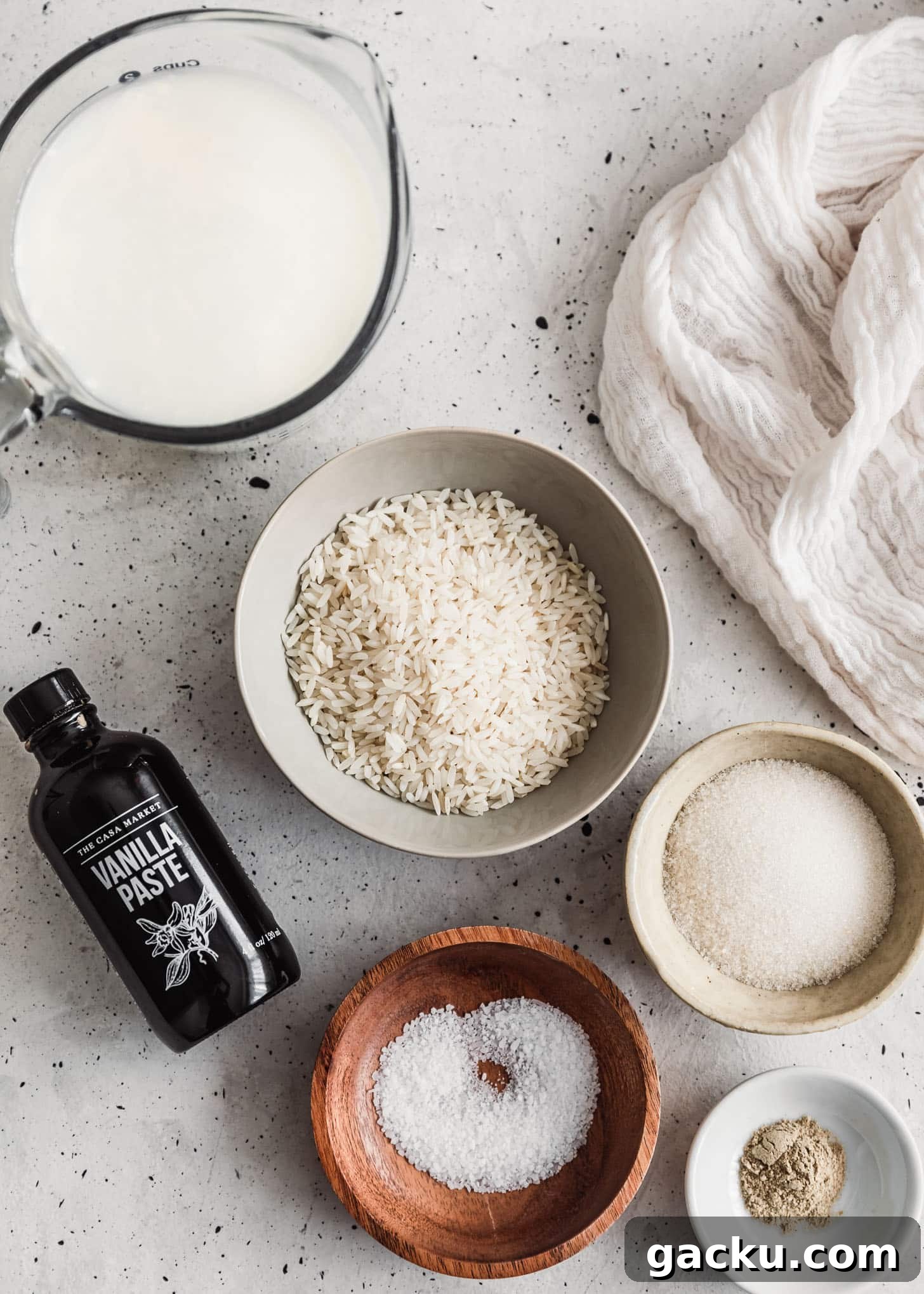 Creamy Cardamom Rice Pudding Recipe — Silky Indian Kheer Guide 3 Wood and white bowls of rice, sugar, milk, cardamom, and salt on a white counter next to a bottle of vanilla bean paste.